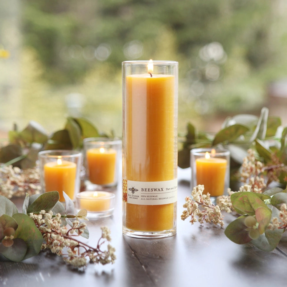 Tall Sanctuary glass beeswax candle lit surrounded by small votives and green foliage on wooden table