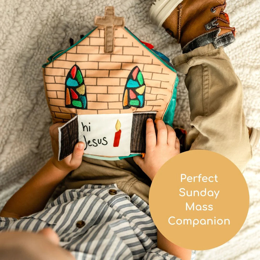 Soft fabric Catholic church-shaped quiet book held by child wearing striped shirt and tan pants on cream blanket