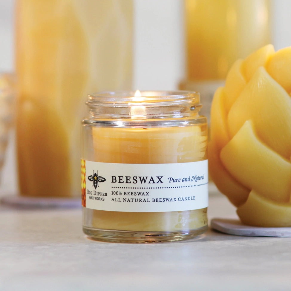 Lit 100% pure beeswax prayer glass candle in clear jar with white label and bee logo on gray surface