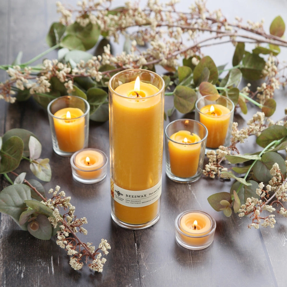 Tall Sanctuary glass beeswax candle surrounded by smaller lit beeswax candles and dried greenery on wood table