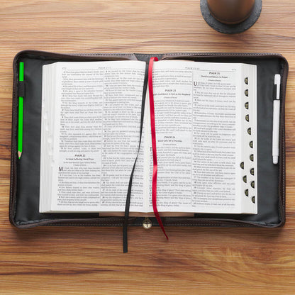 Open Bible inside dark brown leather cover with pen slots, red and black ribbon bookmarks on wooden table