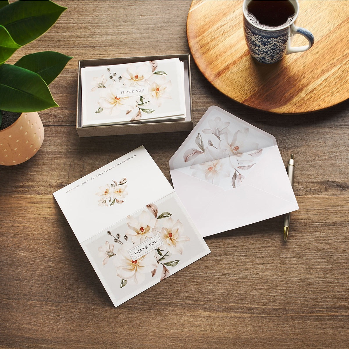 Set of white magnolia thank you note cards with floral design, matching envelopes, and subtle leafy accents on wooden table