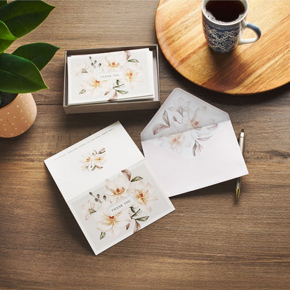 Set of white magnolia thank you note cards with floral design, matching envelopes, and subtle leafy accents on wooden table