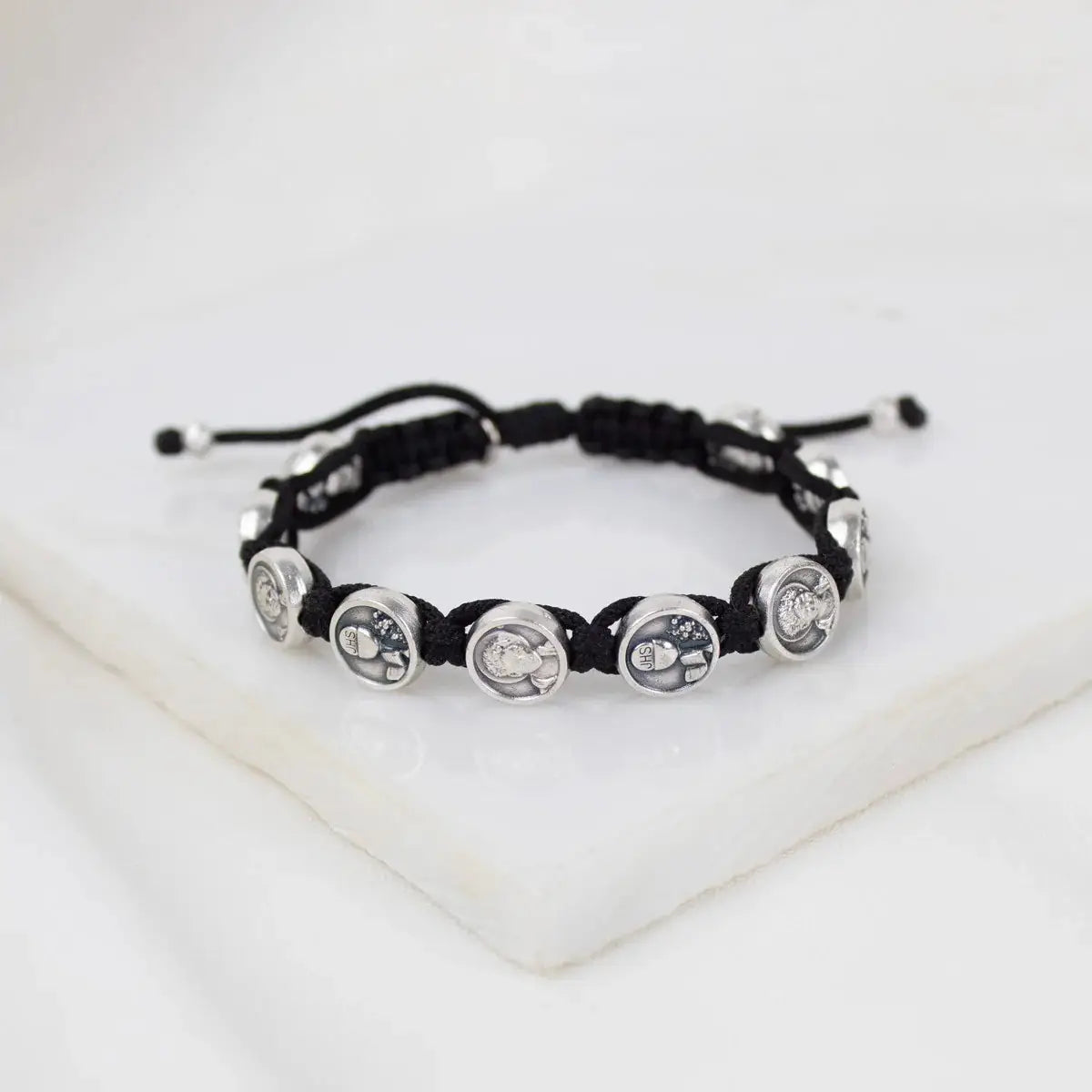 Black cord bracelet with silver Carlo Acutis medals intricately linked, displayed on white marble surface