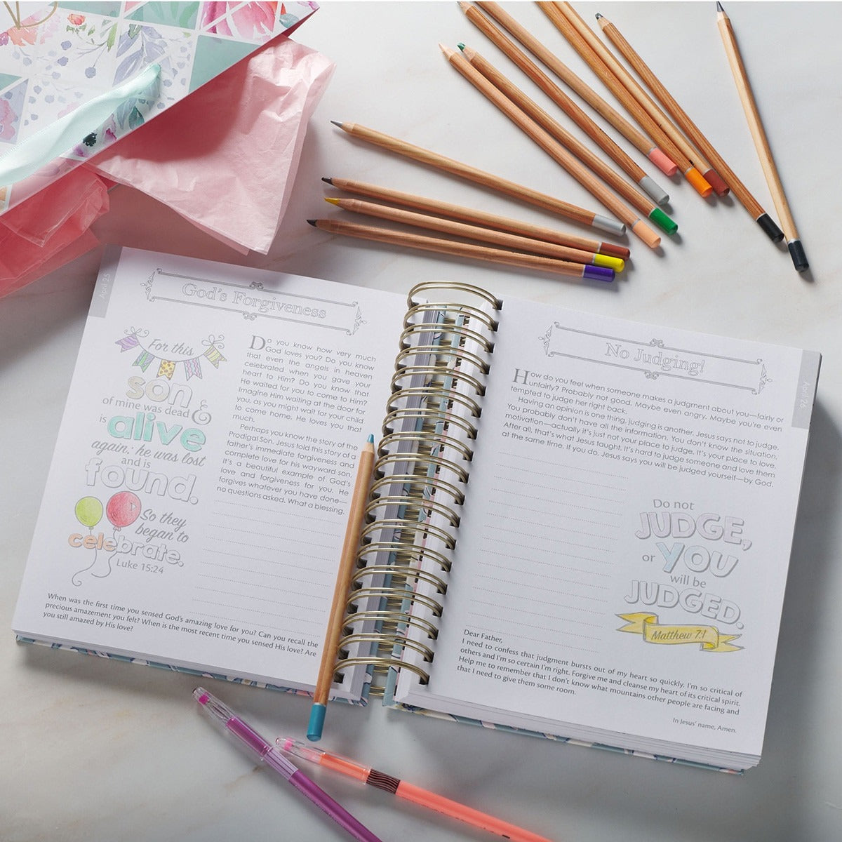 Wirebound devotional coloring book opened to illustrated scripture pages with colored pencils and pens nearby on marble surface