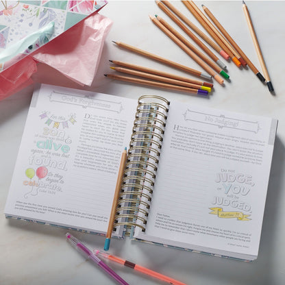 Wirebound devotional coloring book opened to illustrated scripture pages with colored pencils and pens nearby on marble surface