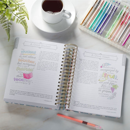 Open wirebound devotional with colorful illustrated words of Jesus, gel pens, coffee cup, and green plant on white table