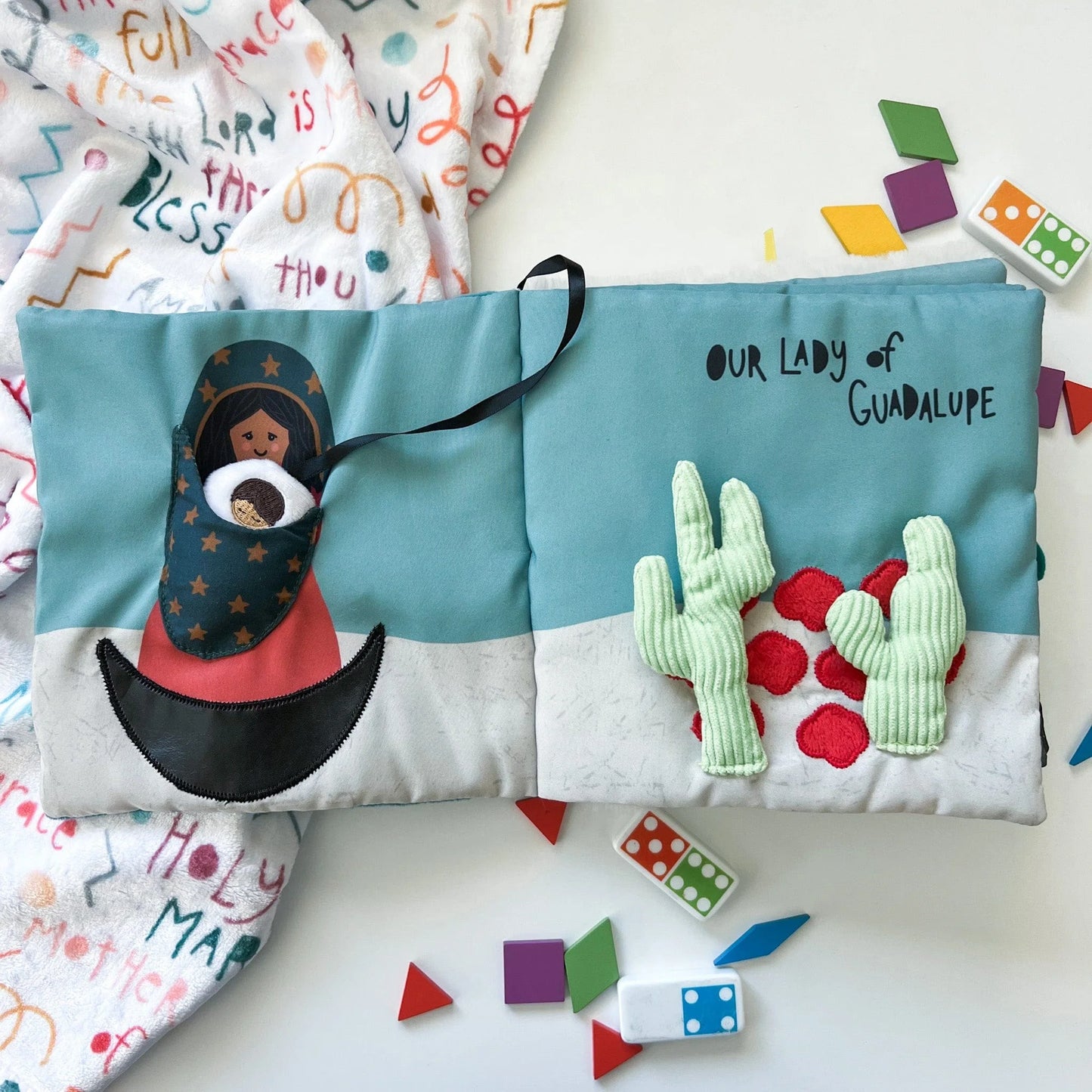 Soft fabric quiet book open to Our Lady of Guadalupe page with embroidered cactus and colorful shapes scattered nearby