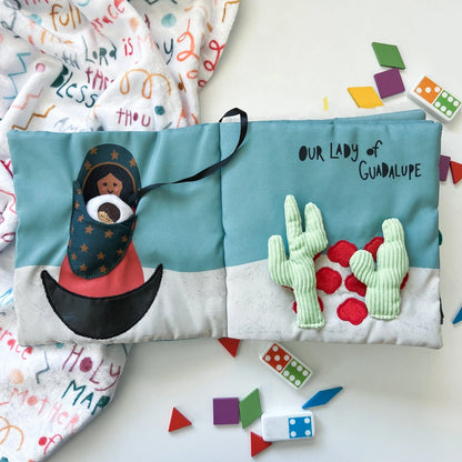 Soft fabric quiet book open to Our Lady of Guadalupe page with embroidered cactus and colorful shapes scattered nearby