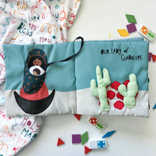 Soft fabric quiet book open to Our Lady of Guadalupe page with embroidered cactus and colorful shapes scattered nearby