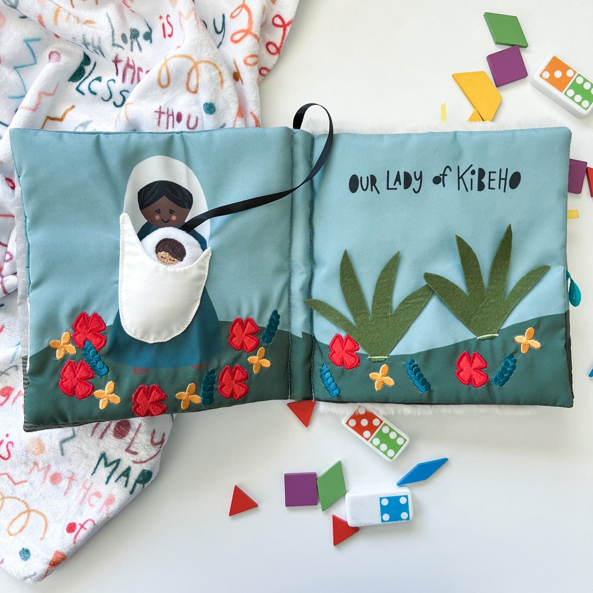 Soft fabric quiet book spread open to reveal embroidered Our Lady of Kibeho with flowers and greenery on blue background