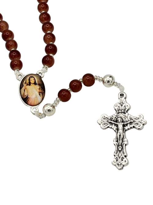 A rosary necklace featuring round reddish-brown beads, silver-tone metal spacers, a detailed silver crucifix, and a central oval medallion with a religious image, displayed on a light gray background.
