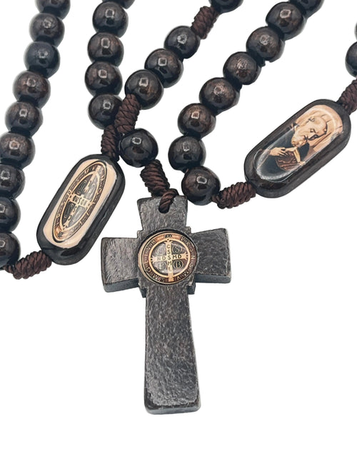 A close-up of a rosary featuring dark brown wooden beads, a dark gray textured cross, and two oval medallions with religious portraits and a central gold-toned emblem, laid on a plain white surface.