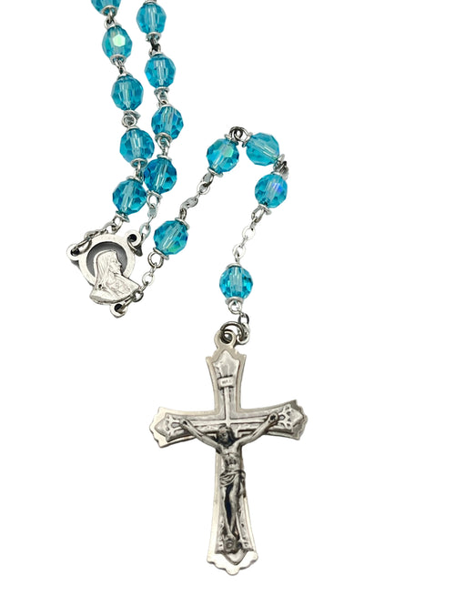 A rosary necklace featuring faceted light blue glass beads and silver-tone metal links, with a silver-tone crucifix pendant and a small silver-tone Our Lady of Mount Carmel medal, displayed on a light gray background.