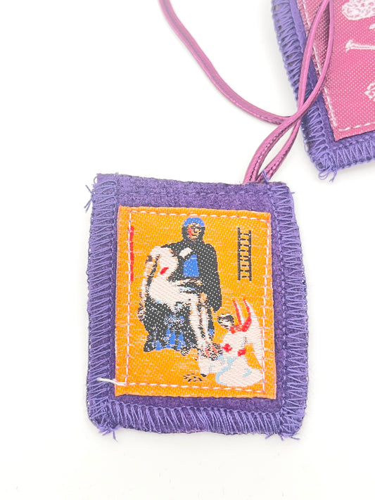 Purple scapular with stitched orange center image of religious figures and purple fabric border, 2 by 1 3/4 inches.