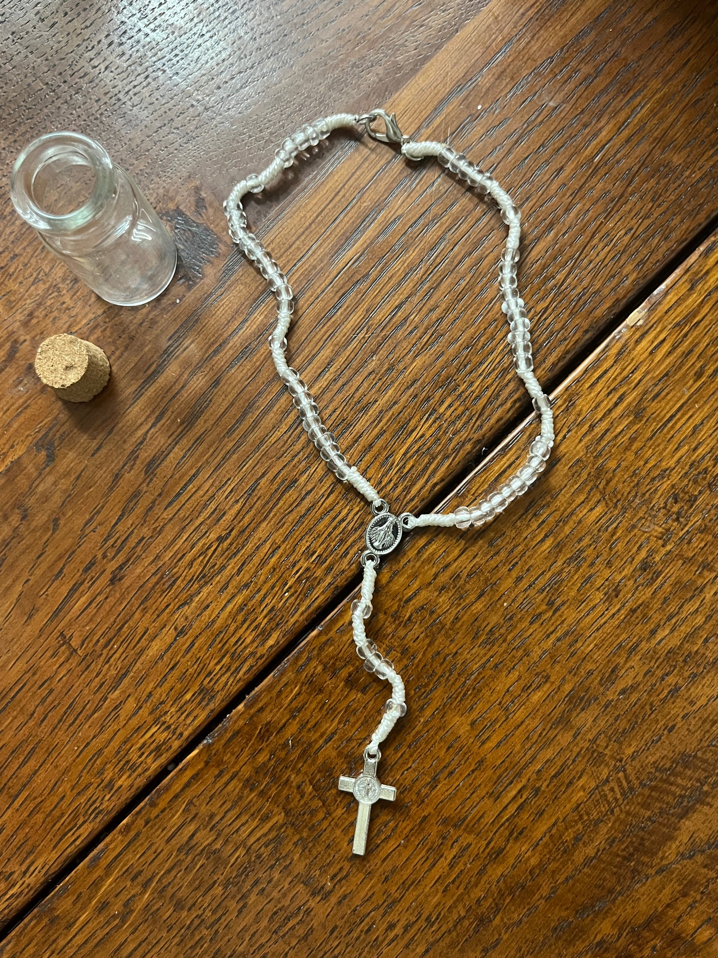 White and clear bead rosary with silver cross and medal, displayed next to empty small glass bottle and cork stopper on wood background.