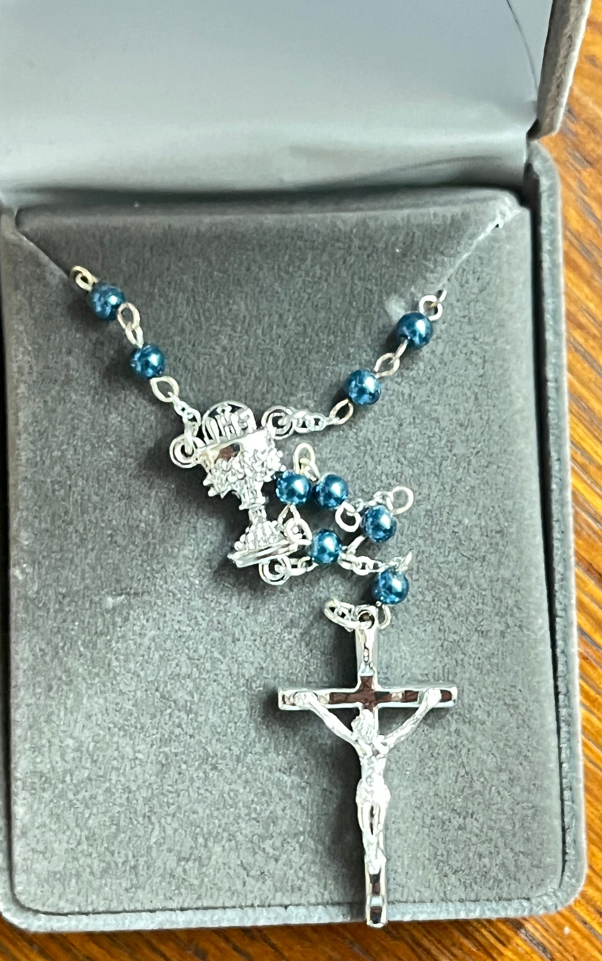 Silver-tone rosary with blue glass beads and chalice centerpiece displayed in gray cushioned box for Communion gift.