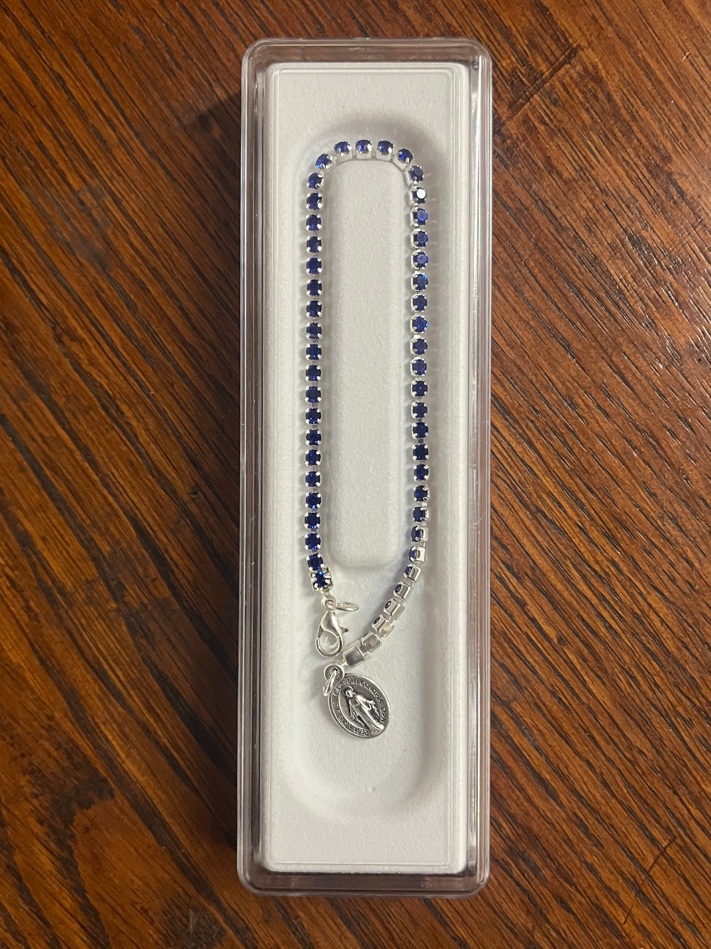 Silver tennis bracelet with blue crystals, Miraculous medal charm, and lobster claw clasp in clear display case