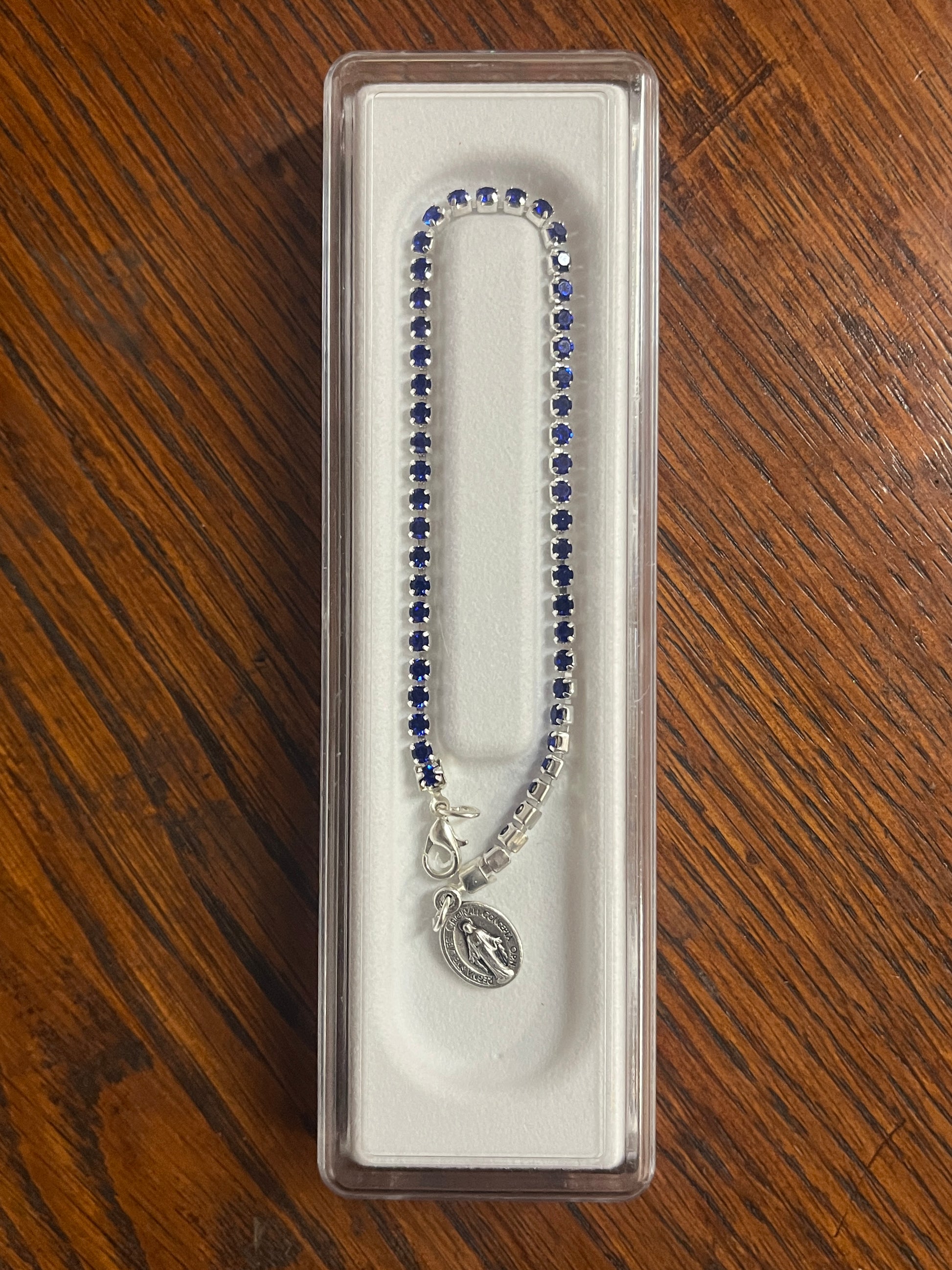 Silver tennis bracelet with blue crystals, Miraculous medal charm, and lobster claw clasp in clear display case
