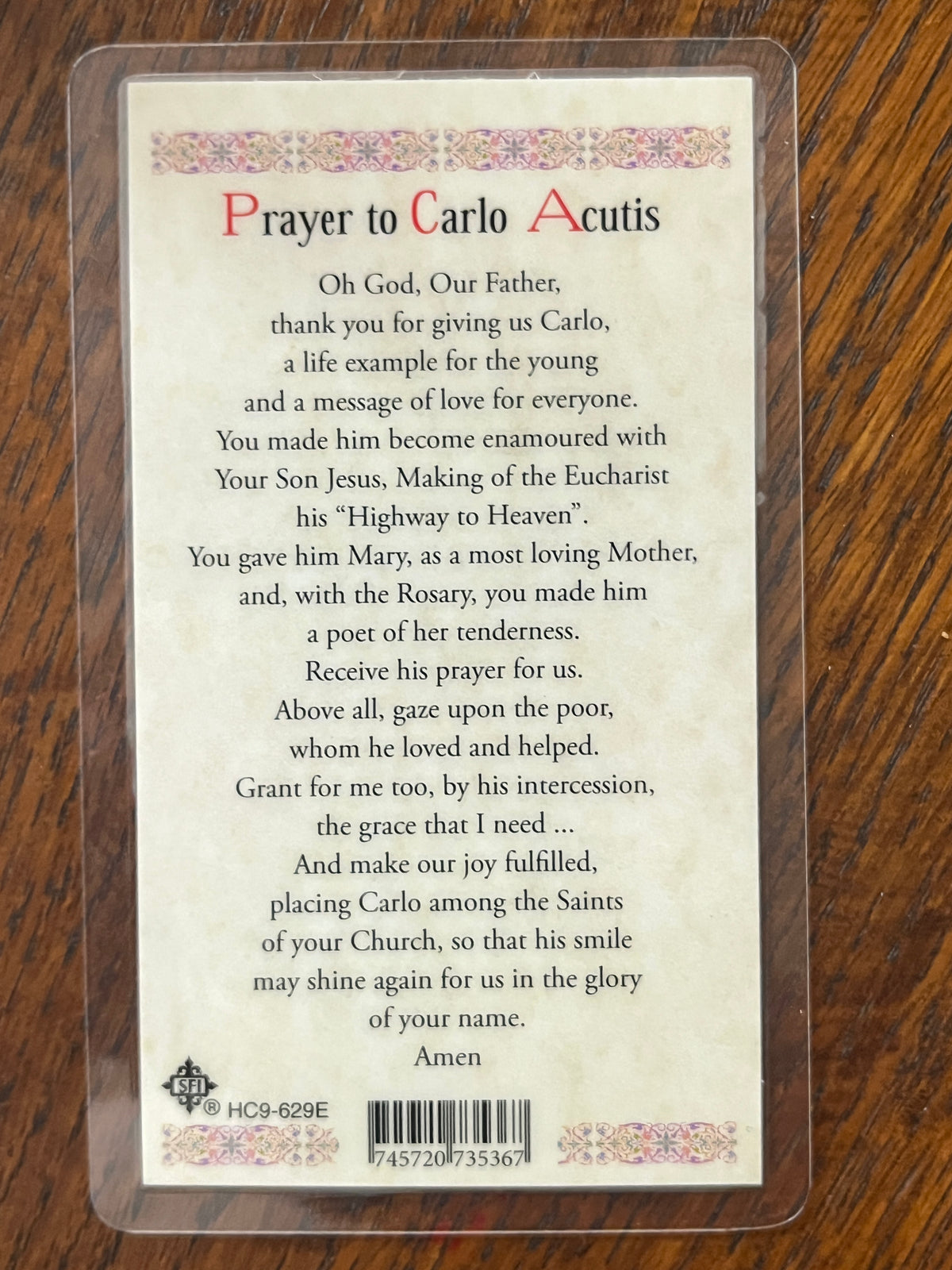 Laminated Holy Card with prayer to St. Carlo Acutis, featuring decorative border and barcode on wooden surface