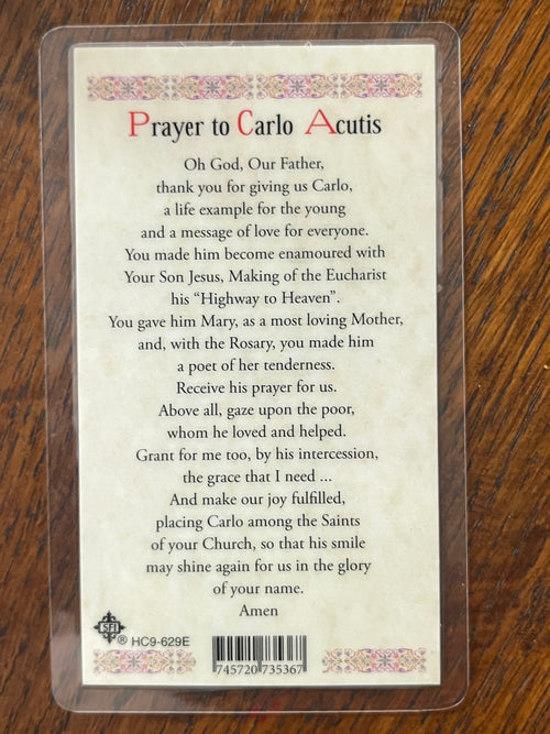 Laminated Holy Card with prayer to St. Carlo Acutis, featuring decorative border and barcode on wooden surface