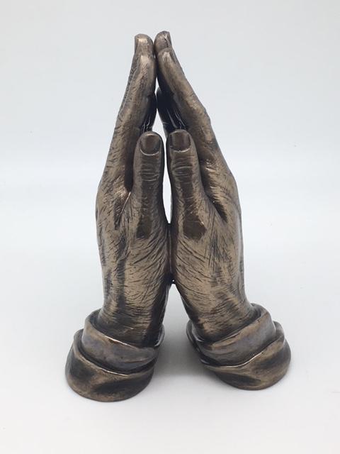 Bronze sculpture of two hands pressed together in prayer with detailed wrist cuffs, standing upright on white background