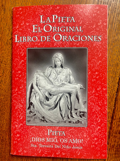 Red cover of La Pieta El Original Libro de Oraciones prayer book with black and white Pieta sculpture image on wood surface