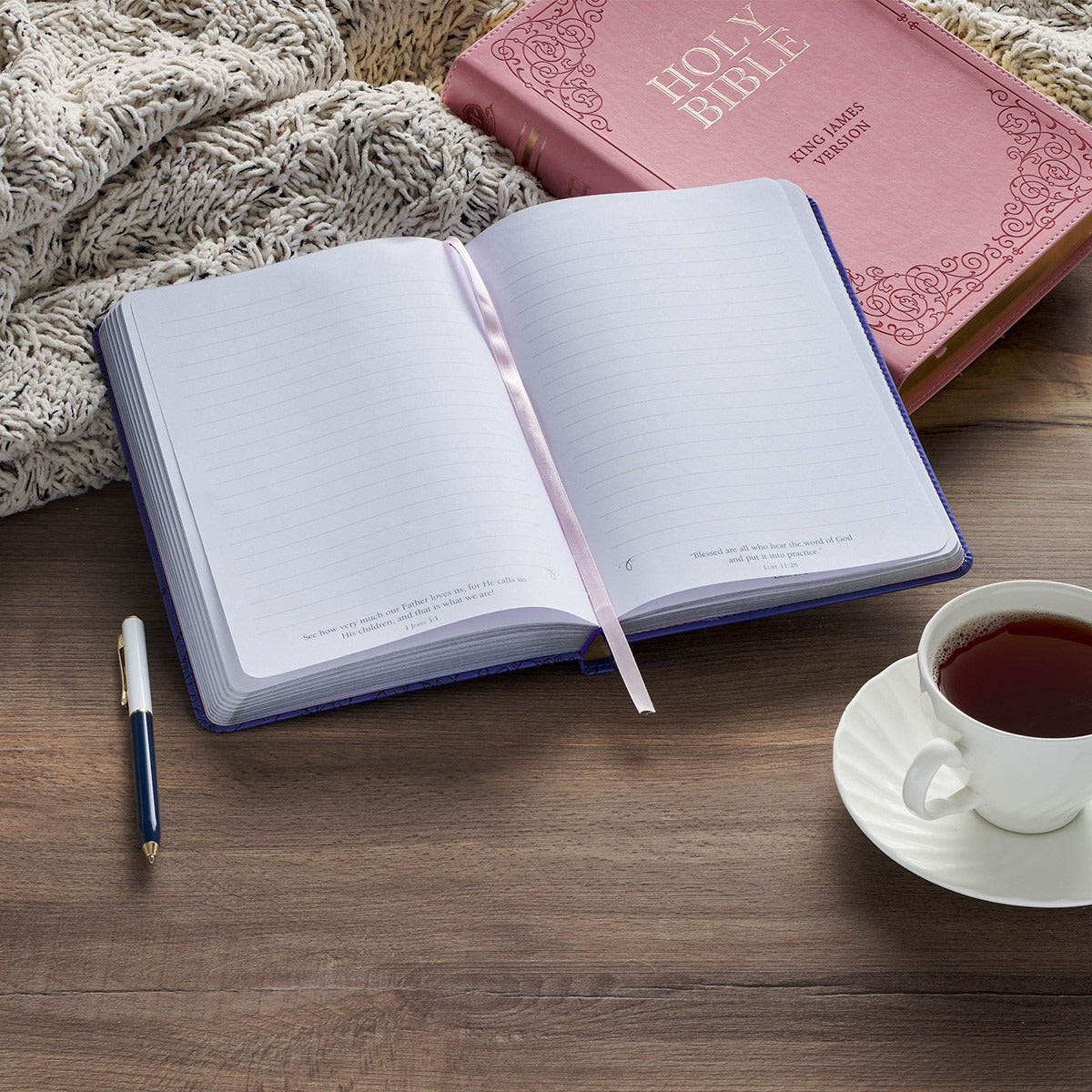 Purple faux leather journal open to lined pages with ribbon marker, pen beside, pink Bible and tea cup on wooden table