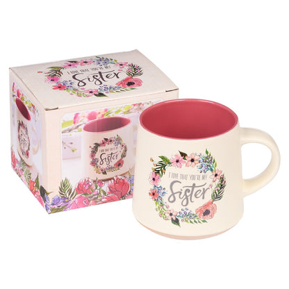 Ceramic coffee mug with pink interior and floral wreath design reading “I love that you’re my sister” with gift box