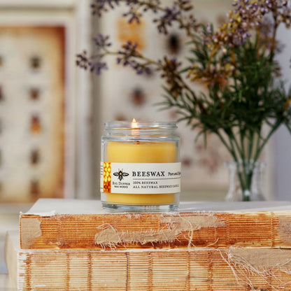 Lit beeswax glass candle with natural honeycomb label on stacked vintage books and blurred flowers behind